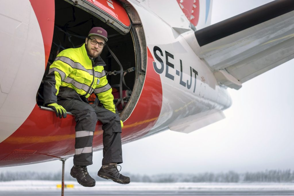 Kertaiseen takkiin ja harmaisiin housuihin pukeutunut mies istuu lentokoneen rahtiluukun laidalla. Jalat roikkuvat luukun ulkopuolella. Lentokone on puna-valkoinen, jonka kyljessä on kirjaimet SE-LJT.