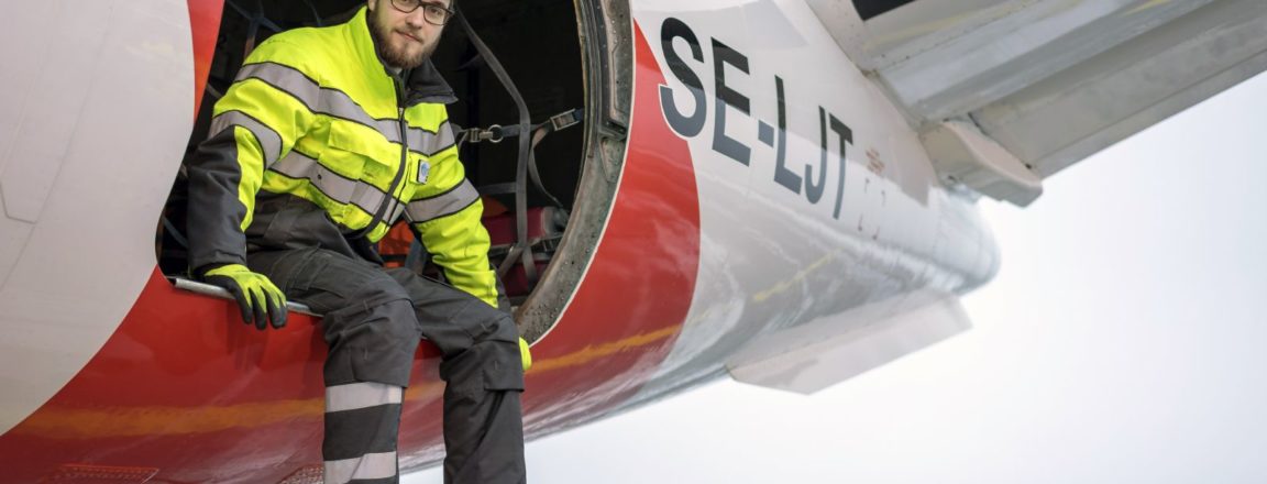 Kertaiseen takkiin ja harmaisiin housuihin pukeutunut mies istuu lentokoneen rahtiluukun laidalla. Jalat roikkuvat luukun ulkopuolella. Lentokone on puna-valkoinen, jonka kyljessä on kirjaimet SE-LJT.