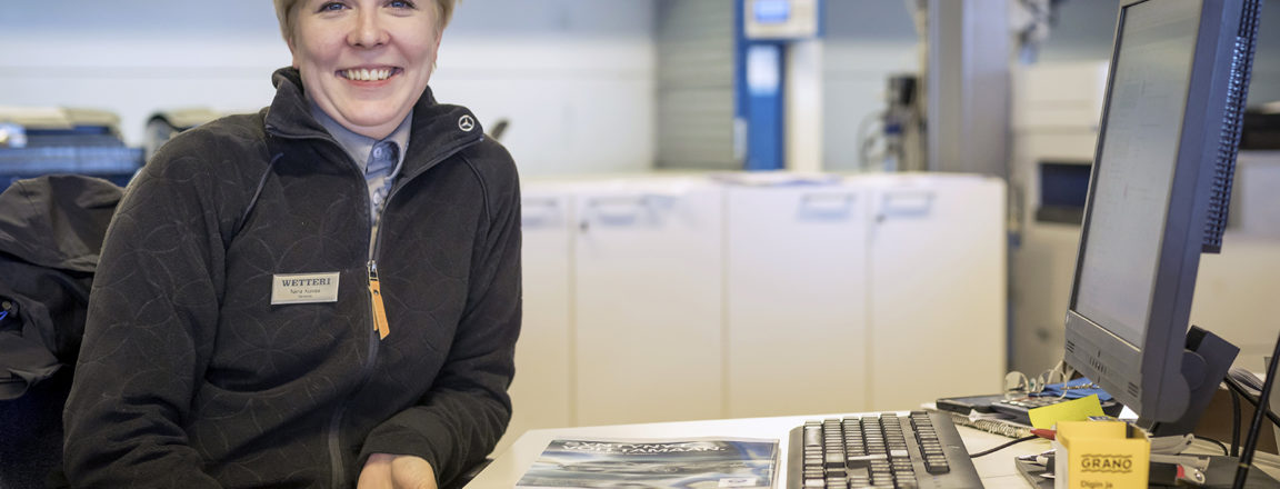 Vaaleahiuksinen nainen istuu työpöydän ääressä. Naisella on päällään tumma fleecetkki, jonka kauluksessa on Mercedes-Bench automerkin logo. Naisen toisessa rinnassa on rintaneula, jossa lukee Wetteri. Pöydällä on tietokone, näppäimistö, hiiri sekä autolehti ja maksupääte.