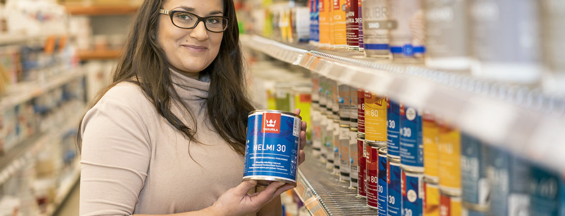 Tummahiuksinen ja silmälasipäinen vaalean roosaan neulepaitaan pukeutunut nainen seisoo rautakaupan maalihyllyllä. Hän pitelee käsissään pientä, litran vetoista metallista maalipurkkia.