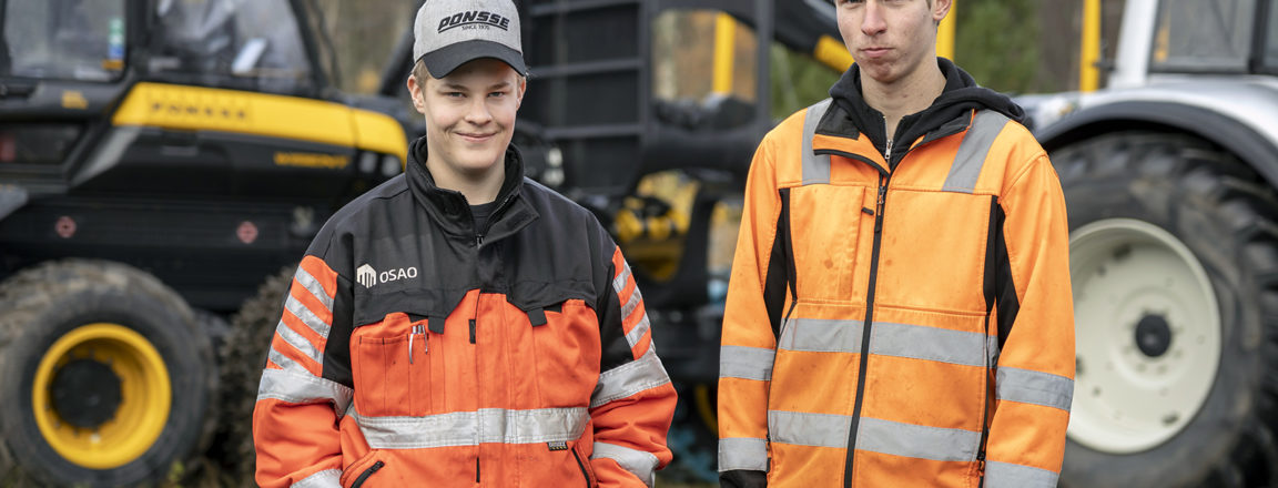 Kaksi oransseihin työtakkeihin pukeutunutta lippalakkipäistä nuorta miestä seisoo kädet taskuissa. Heidän takanaan on kelta-musta metsäkone ja harmaa traktori.