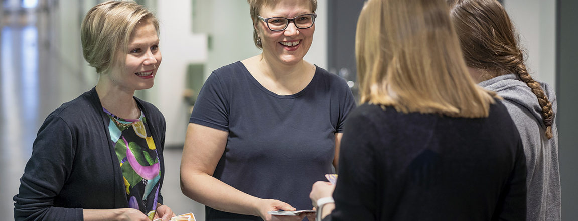 Neljä henkilöä, joista kaksi seisoo selin kameraan. Kasvot kameraan päin seisoo kaksi naista, joilla on vaaleat, lyhyet hiukset.