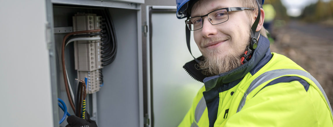 Silmälasipäinen mies sinisessä kypärässä ja keltaisessa takissa pitelee käsissään nippua sähkökaapissa olevia sähköjohtoja.