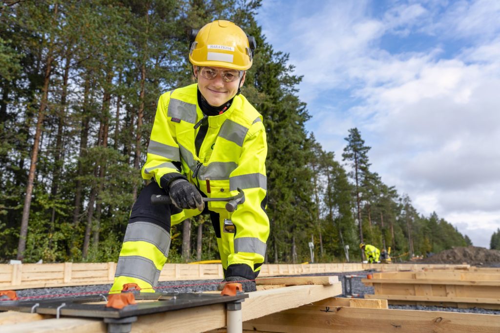 Keltaiseen takkiin ja housuihin sekä keltaiseen kypärään pukeutunut mies seisoo puisen rakennuskehikon keskellä ja pitää vasaraa kädessään. Miehellä on suojalasit päässä ja käsineet kädessä.