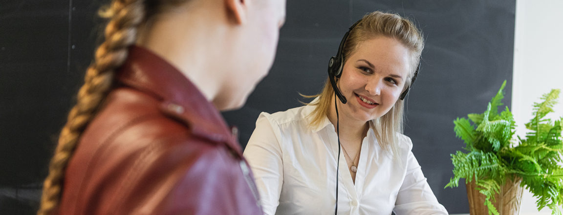 Kaksi naista tutkii pöydällä olevaa paperia. Toisella naista on asiakaspalvelijan headset päässä ja tietokone edessään.