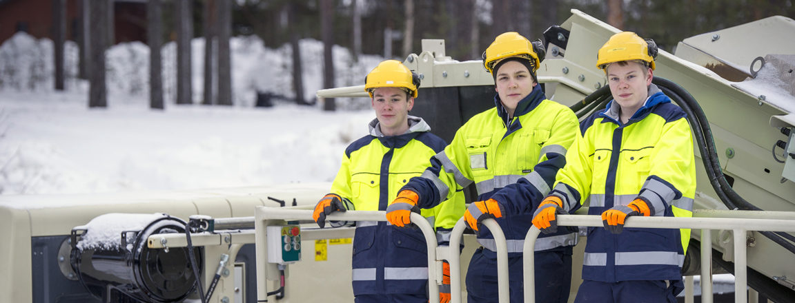 Kolme nuorta miestä keltaisissa turvavaateissa seisovat murskaimen ylätasanteella ja katsovat kohti kameraa. Takana näkyy luminen metsä.