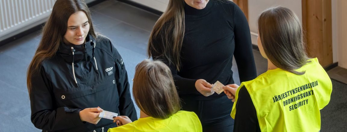 Kaksi keltaiseen liiviin pukeutunutta pitkähiuksista henkilöä tarkistavat edessä olevien naisten henkilökortteja. Keltaisten liivien selässä lukee Järjestyksenvalvoja.