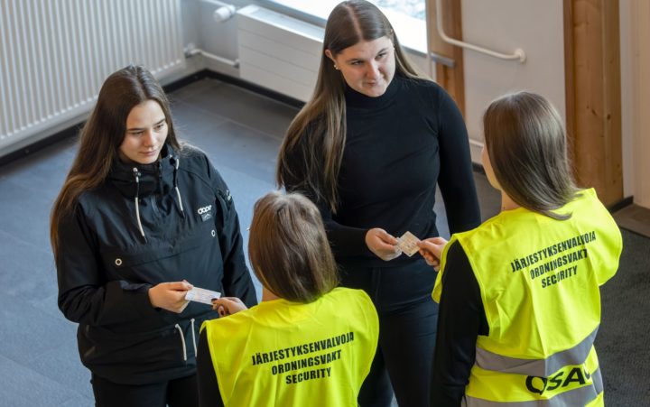 Kaksi keltaiseen liiviin pukeutunutta pitkähiuksista henkilöä tarkistavat edessä olevien naisten henkilökortteja. Keltaisten liivien selässä lukee Järjestyksenvalvoja.