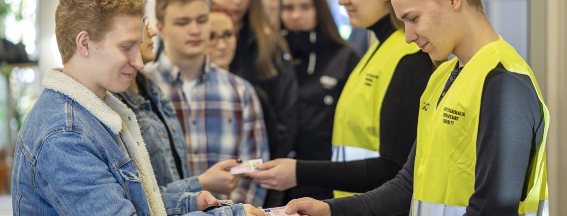 Keltaisiin järjestyksenvalvojan liiveihin pukeutuneet mies ja nainen seisovat ovella ja tarkistavan jonossa olevien henkilöiden henkilökortteja.