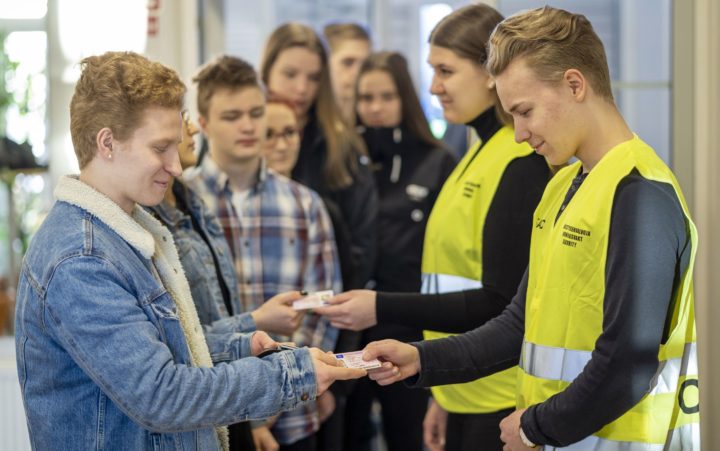 Keltaisiin järjestyksenvalvojan liiveihin pukeutuneet mies ja nainen seisovat ovella ja tarkistavan jonossa olevien henkilöiden henkilökortteja.