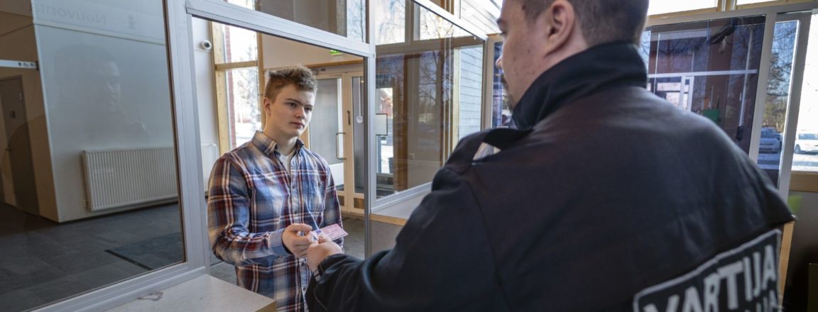 Tummahiuksinen, tummaan haalariin pukeutunut mies, jonka haalarin selässä lukee VARTIJA-opiskelija, seisoo puisen tiskin takana. Mies ojentaa lasiseinän toisella puolella seisovalle ruutupaitaiselle miehelle henkilökorttia.