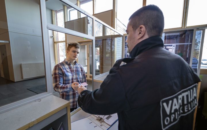 Tummahiuksinen, tummaan haalariin pukeutunut mies, jonka haalarin selässä lukee VARTIJA-opiskelija, seisoo puisen tiskin takana. Mies ojentaa lasiseinän toisella puolella seisovalle ruutupaitaiselle miehelle henkilökorttia.