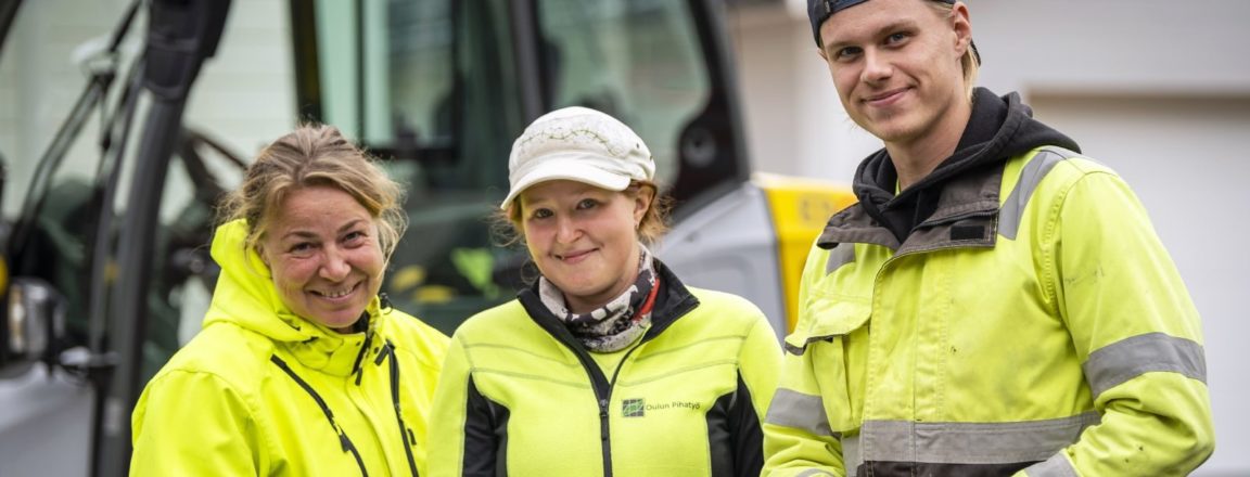 Kolme keltaisiin takkeihin pukeutunutta henkilöä seisoo traktoria muistuttavan koneen edessä. Kaksi vasemmanpuoleista henkilöä on naisia ja oikeanpuoleisin mies. Miehellä on kädessään lapio ja keskimmäinen henkilöistä pitää käsissään piirustusta.