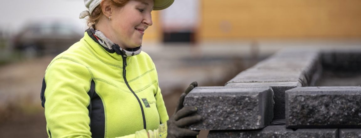 Lippalakkiin ja keltaiseen takkiin pukeutunut nainen asettelee kiveä paikalleen rakentamaansa kivimuuriin. Takana näkyy valmistumassa oleva oranssiin vivahtava puutalo, jonka pihalla on multaa.