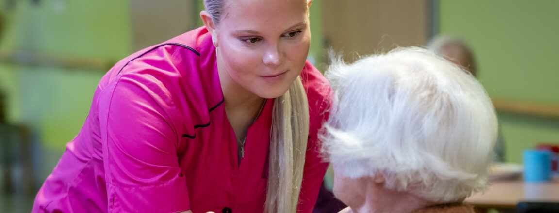 Pinkkiin asuun pukeutunut hoitaja jututtaa vanhusta.