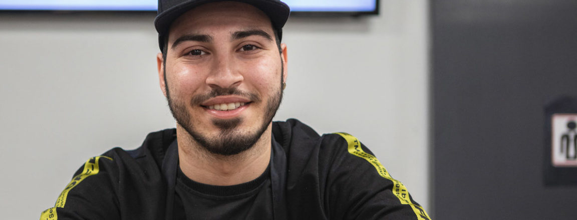 The young man is wearing a black shirt and cap. He leans with his arms on the glass counter.