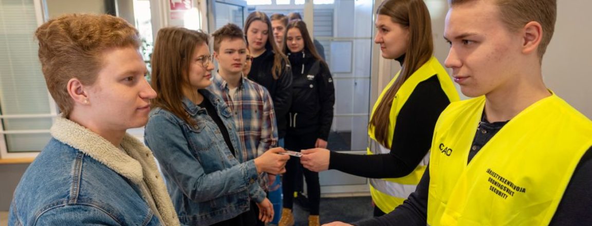 Keltaisiin järjestyksenvalvojan liiveihin pukeutuneet mies ja nainen seisovat ovella ja tarkistavan jonossa olevien henkilöiden henkilökortteja.