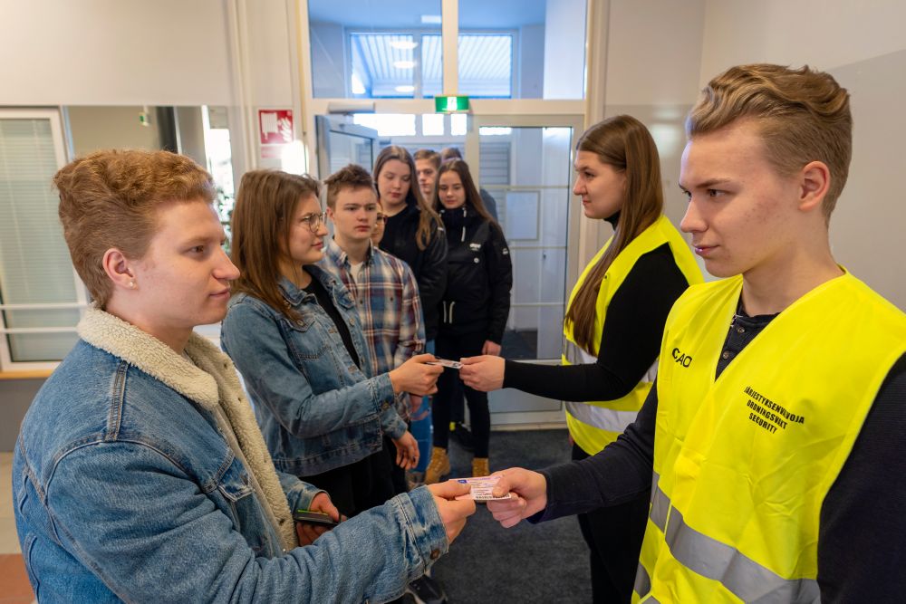 Keltaisiin järjestyksenvalvojan liiveihin pukeutuneet mies ja nainen seisovat ovella ja tarkistavan jonossa olevien henkilöiden henkilökortteja.