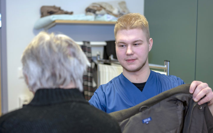 Nuori mies hoitajan sinisessä työasussa auttaa takkia ikääntyneen henkilön ylle.