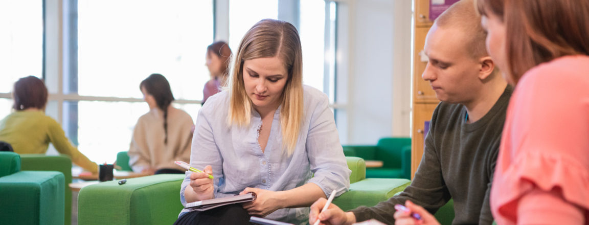 Vaaleahiuksinen nainen kirjoittaa vihkoon, vaaleahiuksinen mies osoittaa jotain tabletistaan ja punertavahiuksinen nainen katsoo tablettia. Naiset ja mies istuvat vihreillä sohvilla. Taustalla hylly sekä muita ihmisiä.