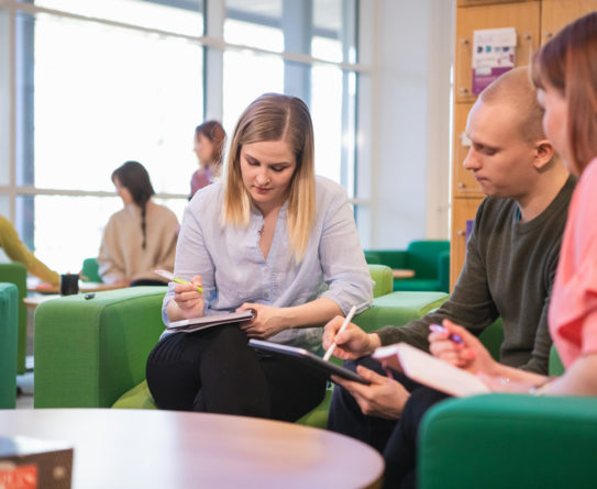 Vaaleahiuksinen nainen kirjoittaa vihkoon, vaaleahiuksinen mies osoittaa jotain tabletistaan ja punertavahiuksinen nainen katsoo tablettia. Naiset ja mies istuvat vihreillä sohvilla. Taustalla hylly sekä muita ihmisiä.