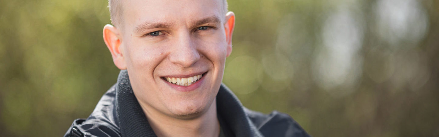 Short-haired man looking and smiling at the camera. There's a greenery behind him.