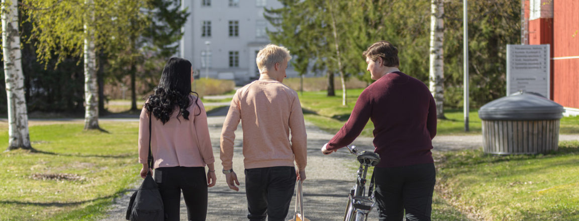 Kolme henkilöä kävelee pihatietä pitkin kohti valkoista isoa rakennusta. Oikeanpuoleinen henkilöistä taluttaa polkupyörää.