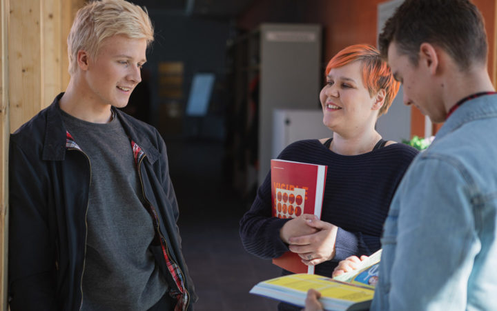 A woman and two men stand and talk to each other. She has a book in her hand.