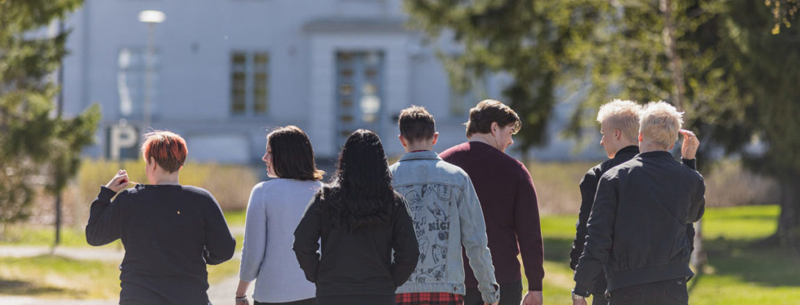 A group of students are walking away from the camera towards a white building. There's greenery by the side.