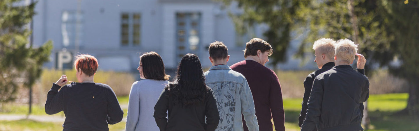 A group of students are walking away from the camera towards a white building. There's greenery by the side.