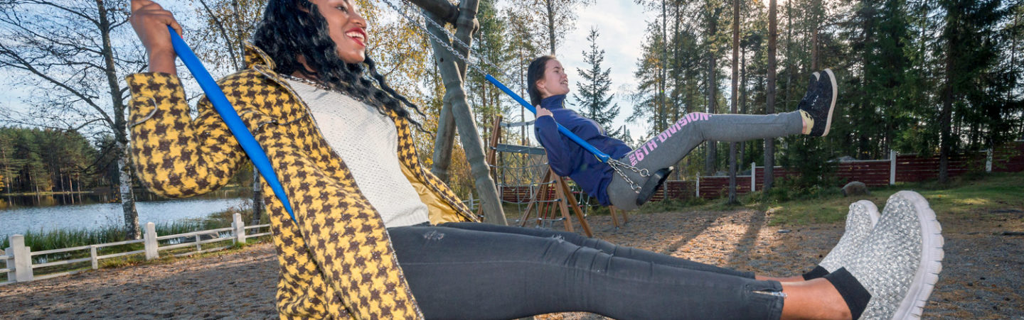 Two dark-haired young women are swinging outside. There's sand on the ground and the sun is shining between the trees.