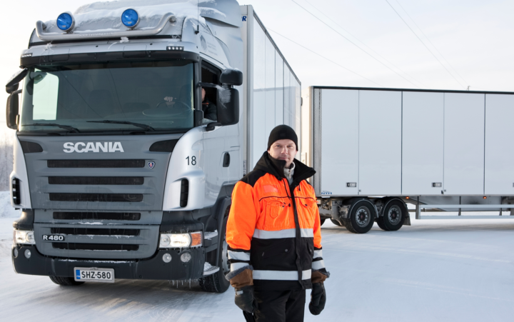 Mies, jolla on oranssi huomiotakki päällään seisoo valkoisen Scania-merkkisen yhdistelmäajoneuvon edessä. Maassa on lunta ja miehellä pipo päässä.