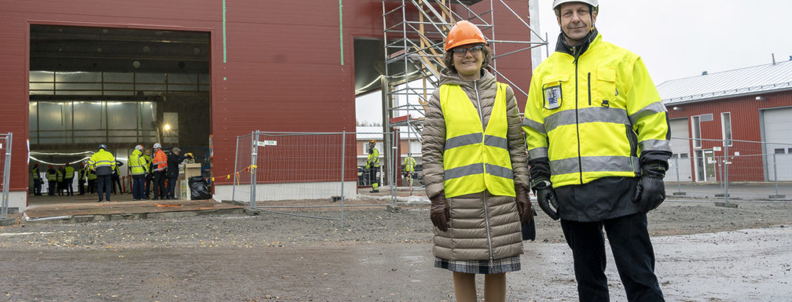 Huomioliiviin pukeutunut oranssikypäräpäinen nainen ja huomiotakkiin pukeutunut valkokypäräinen mies seisovat rakenteilla olevan punaisen hallirakennuksen edessä.