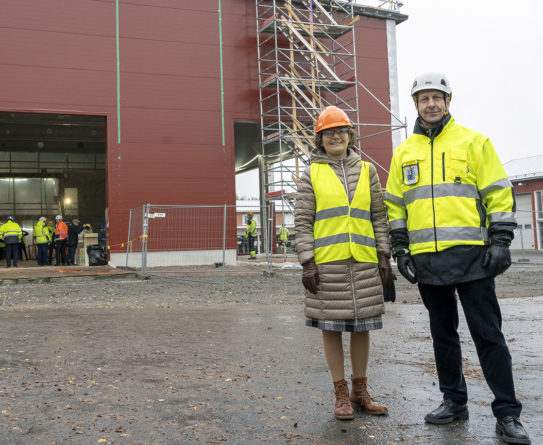 Huomioliiviin pukeutunut oranssikypäräpäinen nainen ja huomiotakkiin pukeutunut valkokypäräinen mies seisovat rakenteilla olevan punaisen hallirakennuksen edessä.