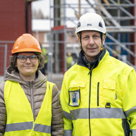 Huomioliiviin pukeutunut oranssikypäräpäinen nainen ja huomiotakkiin pukeutunut valkokypäräinen mies seisovat rakenteilla olevan punaisen hallirakennuksen edessä.