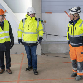 Kolme miestä keskustelee rakenteilla olevassa rakennuksessa. Vasemmalla olevalla miehellä on oranssi kypärä ja keltainen huomioliivi. Keskellä olevan mihen päässä on valkoinen kypärä, päällä keltainen huomiotakki ja jalassa farkut. Oikeanpuoleisella miehellä on valkoinen kypärä, keltainen huomiotakki ja oranssit huomionväriset työhousut.