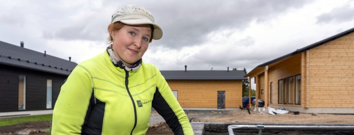 Lippalakkiin ja keltaiseen takkiin sekä käsineisiin pukeutunut nainen asettelee kiveä paikalleen rakentamaansa muuriin. Muurin toiseen reunaan on tehty istutuksille koko muurin mittainen kaukalo, jossa on istutuksia varten multaa. Takana näkyy oranssiin vivahtava valmistumassa oleva puutalo, jonka pihalle on levitetty mullat.