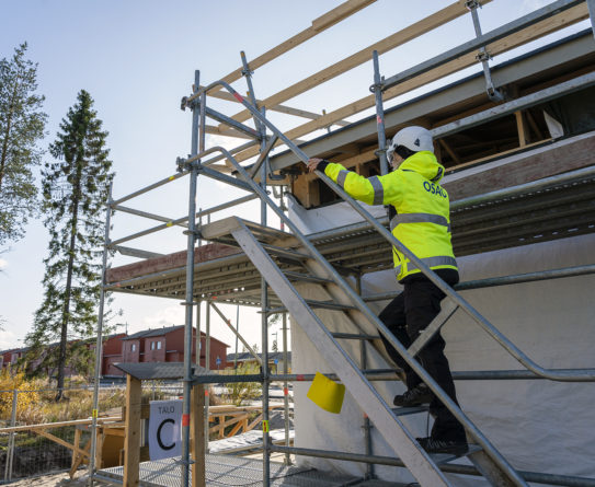 Keltatakkinen henkilö kiipeää rakennuksen yhteydessä olevilla rakennustelineillä. Henkilöllä on päässä valkoinen kypärä. Taustalla näkyy toinen rakennus ja puu.