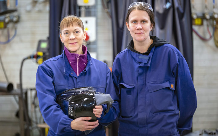 Kaksi naista seisoo vierekkäin työsalissa, sinisiin työhaalareihin pukeutuneina. Vasemman puoleinen pitää käsissään hitsauskypärää. Taustalla työsalin varustusta.