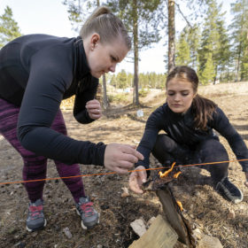 Kaksi naista on kyykyssä ja puhaltaa pienessä nuotiossa olevaa tulta. Nuotion yläpuolelle on pingotettu punainen lanka.