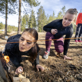 Kaksi naista on kyykyssä ja puhaltaa pienessä nuotiossa olevaa tulta. Nuotion yläpuolelle on pingotettu punainen lanka.