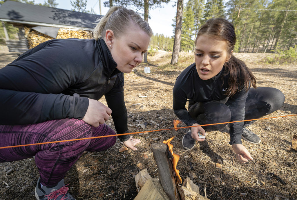 Kaksi naista on kyykyssä ja puhaltaa pienessä nuotiossa olevaa tulta. Nuotion yläpuolelle on pingotettu punainen lanka.