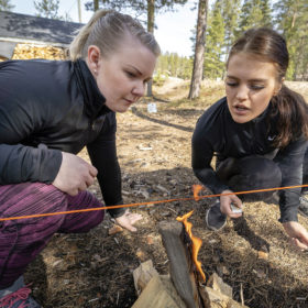 Kaksi naista on kyykyssä ja puhaltaa pienessä nuotiossa olevaa tulta. Nuotion yläpuolelle on pingotettu punainen lanka.