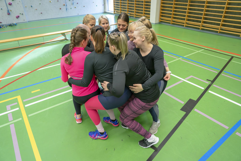 Joukko liikuntavaatteisiin pukeutuneita henkilöitä seisoo tiiviissä kasassa keskellä liikuntasalia. Näyttää, että henkilöt ovat istumassa toistensa syleihin.