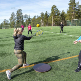 Joukko henkilöitä on urheilukentällä ja pelaa pesäpalloa tennismailalla ja keltaisella pallolla.