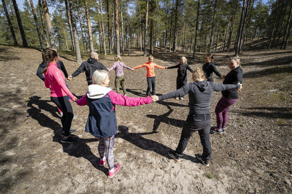 Joukko liikunnallisiin vaatteisiin pukeutuneita henkilöitä seisoo mäntymetsässä piirissä ja pitää toisiaan kädestä kiinni.