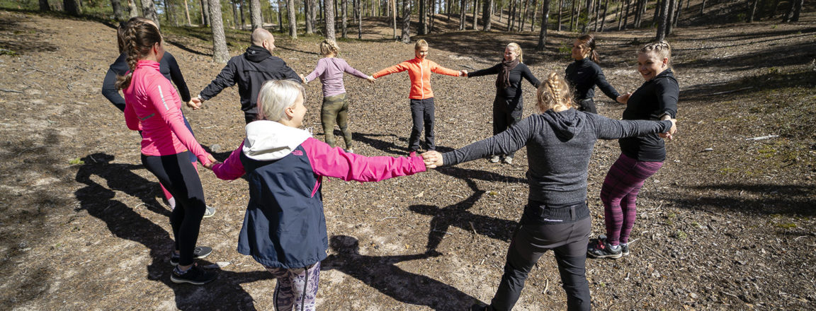 Joukko liikunnallisiin vaatteisiin pukeutuneita henkilöitä seisoo mäntymetsässä piirissä ja pitää toisiaan kädestä kiinni.