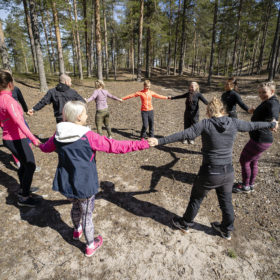 Joukko liikunnallisiin vaatteisiin pukeutuneita henkilöitä seisoo mäntymetsässä piirissä ja pitää toisiaan kädestä kiinni.