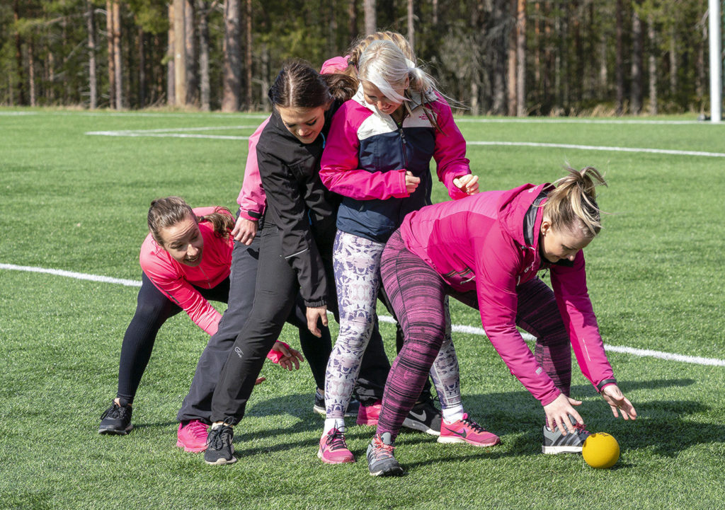 Viisi naista on jonossa. Jonon ensimmäinen on pukkaamassa keltaista palloa takana olevien haarojen välistä. Jonon takimmainen valmistautuu ottamaan pallon vastaan.