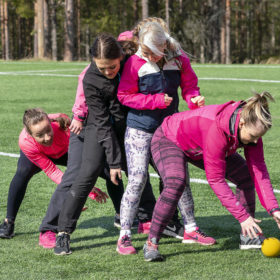 Viisi naista on jonossa. Jonon ensimmäinen on pukkaamassa keltaista palloa takana olevien haarojen välistä. Jonon takimmainen valmistautuu ottamaan pallon vastaan.